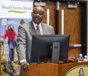 A man in a suit speaks at a podium with a computer, in front of a Small Farms Week banner. Behind him, a blurred background shows a wooden wall and a door. Two water bottles are on the podium.