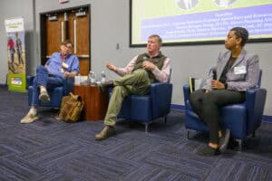 Three panelists are seated on stage in front of a screen. The middle panelist is speaking, gesturing with one hand. They sit on either side of a small table with water bottles. The background features a banner and a PowerPoint presentation.
