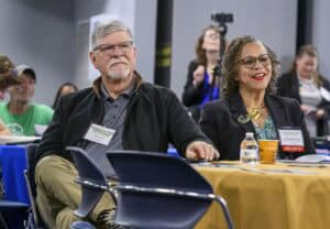Two people sit at a table during a conference. Both wear conference badges. The man has gray hair and glasses, and the woman is smiling, wearing glasses and a badge labeled Speaker. Other attendees and a camera on a tripod are visible in the background.