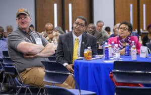 A diverse group of people seated at a table with blue tablecloth, engaged in a presentation. Three individuals in the foreground, including a man in a cap and two in business attire, listen attentively. Bottles and snacks are on the table.