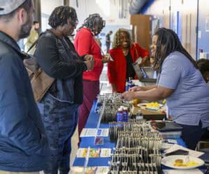 People gather around a table at an event. A woman in a blue shirt is distributing items from the table, which is lined with pens, brochures, and snacks. Other attendees, including two women, are engaged in the event activities.