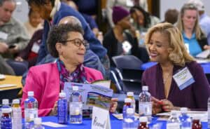 Two women seated at a table during an event. One, wearing a pink jacket, holds a pamphlet about Small Farmers. The other, in a dark purple jacket, is smiling. Bottles of water and name tags are visible. Attendees are in the background.