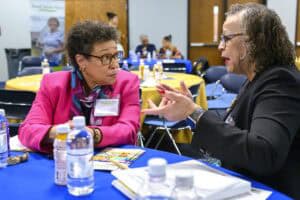 Two women are seated at a table with a blue tablecloth, engaged in conversation. The table has water bottles and documents. Both women are wearing glasses, and people are visible in the background in a conference setting.