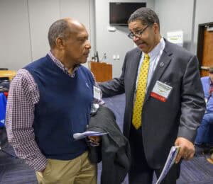 Two men are having a conversation in a conference room. One is holding a coat and a notebook, while the other gestures with one hand. They are dressed in business attire and wearing conference badges.