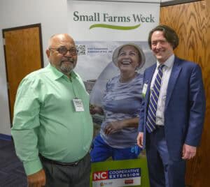 Two men stand in front of a Small Farms Week banner. One man is wearing a green shirt and glasses, the other is in a blue suit and tie. The banner includes an image of a smiling person in a garden.