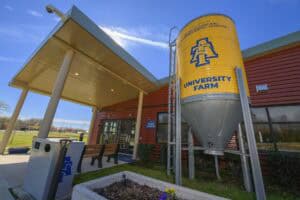 Entrance of a red farm building with a tall, yellow silo labeled University Farm and university logo. A bench and trash cans are in the foreground, and blue sky with a visible contrail is overhead.