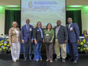 A group of six people stand together, smiling, in front of a screen displaying North Carolina Agricultural and Technical State University with a university emblem. One person holds a plaque. They are on a stage with yellow and blue floral arrangements.
