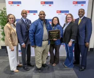 A group of six people stands together, smiling at the camera. One person holds a plaque that reads Small Farms Week. Behind them is a banner featuring the NC Cooperative Extension logo.