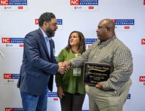 Three people stand in front of NC Cooperative Extension signs. Two men are shaking hands, and one man holds a plaque. A woman stands in the middle, looking at them, smiling.