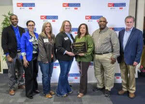 A group of seven people stand together, posing with a plaque in front of a backdrop that reads NC Cooperative Extension. The individuals are dressed in business casual attire, smiling for the photo.