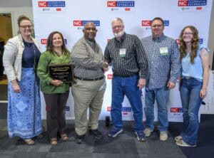 Group of six people standing in front of an NC Cooperative Extension backdrop, with Elvin and Madeline Eaton holding a Small Farms Week plaque; Elvin Eaton shakes hands with another man in a checkered shirt, while others smile and stand on either side in casual or business-casual attire.