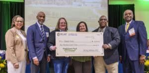 Six people standing on stage in front of a projected farm image, smiling and holding a ceremonial check for $2,000 awarded to Fairport Farms during Small Farms Week; two individuals at center hold the check while others stand on either side, dressed in professional attire.