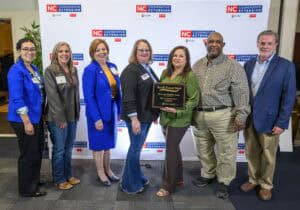 Group of seven people standing in front of an NC Cooperative Extension backdrop, with two individuals in the center holding a Small Farms Week plaque recognizing Elvin and Madeline Eaton as 2025 Small Farmers of the Year; all are smiling and dressed in business or business-casual attire.