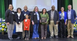 Group of nine people standing on a stage in front of a projected screen and flower arrangements, with two individuals in the center holding a Small Farms Week plaque; all are smiling and wearing business or business-casual attire, including a young girl in a red dress.