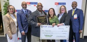 Group of seven people standing in front of a backdrop labeled "NC Cooperative Extension," with the central individual holding a plaque reading "Small Farms Week" and a large ceremonial check for $2,000 made out to Fairport Farm.