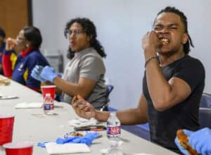 Three people sit at a table eating chicken wings. The person in the foreground, wearing a black shirt, is enthusiastically biting into a wing. Food, napkins, water bottles, and gloves are on the table.