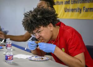 A young person wearing a red shirt, glasses, and blue gloves eats chicken wings at a table with water bottles and a paper plate in front of them. A yellow banner hangs on the wall in the background.