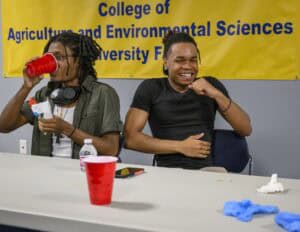 Two young men sit at a table with red cups, a water bottle, and a phone, smiling and laughing. A yellow banner behind them reads College of Agriculture and Environmental Sciences University.