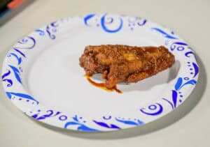 A single sauced chicken wing sits on a white paper plate with blue decorative patterns, placed on a light-colored surface.