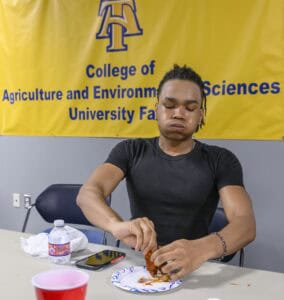 A person with puffed cheeks eats spicy chicken wings at a table, in front of a yellow College of Agriculture and Environmental Sciences banner. A water bottle, phone, and red cup are on the table.