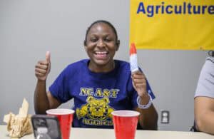 A smiling person wearing an NC A&T Aggies shirt gives a thumbs up while holding a red popsicle. There are red cups on the table and a yellow Agriculture sign in the background.