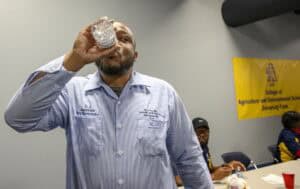 A man in a light blue work shirt drinks from a water bottle in a room. In the background, two people sit at a table near a yellow banner for the College of Agriculture and Environmental Sciences.