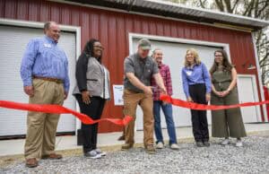 A group of six people stand in front of a red building with two white garage doors. A man in the center cuts a red ribbon, holding large scissors. They are all smiling, and there is gravel on the ground in front of them.