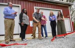 A group of six people stands in front of a red building with two open garage doors. One person is cutting a red ribbon on a gravel path, while the others clap and smile.