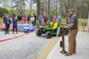 A man in a cap and casual clothing speaks at a microphone on an outdoor stage. A red ribbon is stretched in front of him. Behind, a crowd stands near tables with checkered tablecloths and a small green utility vehicle in a wooded area.