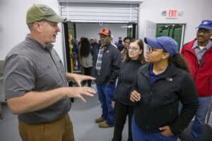 A man in a gray shirt and cap speaks animatedly to a group of people gathered around him in an indoor setting. The group includes two women and several men, some wearing hats and casual clothing, listening attentively. An exit sign is visible in the background.