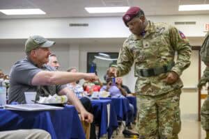A soldier in camouflage fist bumps a seated man wearing a cap inside a room with tables and people. The soldier has numerous patches on his uniform, and both appear engaged in friendly interaction.