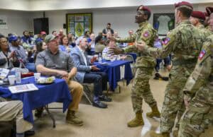 Soldiers in uniform interact with seated attendees at an indoor event. The room has decorated tables, and several people are clapping. Some soldiers wear maroon berets, indicating they might be paratroopers. Walls display various military-themed decorations.