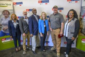 A group of seven people smiling and standing in front of a backdrop featuring NC Cooperative Extension logos. They appear to be dressed for a professional event or gathering.
