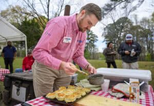 A man in a red checkered shirt applies glaze to biscuits on a tray outdoors. Hes at a table with red-checkered cloth, with others in the background. Trees and a tent are visible. A box of Ziploc bags is on the table.