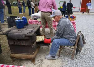 A man in a gray shirt and cap is seated and removing biscuits from a vintage black stove outdoors. He wears a red oven mitt. People are gathered in the background near tables with checkered tablecloths. Gravel covers the ground.