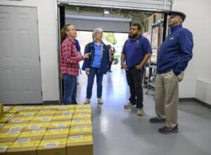 Four people stand in a warehouse near stacks of yellow boxes. Two women, one in a red plaid shirt and another in a blue jacket, converse with two men wearing blue jackets and khaki pants. A partially open garage door is in the background.