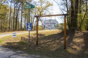 A wooden sign for Secret Gardens Bees & Honey Farm stands beside a road. The sign is mounted on a wooden frame, with street signs for Laurinburg Rd and Moray St nearby. Trees and a blue sky are in the background. A directional sign points left.