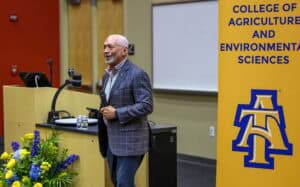 A man in a checked blazer speaks while holding a microphone at a podium, next to a sign reading College of Agriculture and Environmental Sciences with a blue and yellow logo. Yellow flowers are in the foreground.