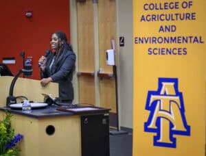 A woman speaks at a podium with a microphone in a lecture hall. Next to her is a yellow banner reading College of Agriculture and Environmental Sciences with a blue and gold logo.