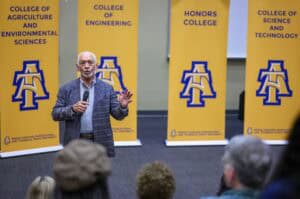 A man speaks into a microphone on stage in front of four yellow banners representing different colleges at North Carolina A&T State University, while an audience listens.