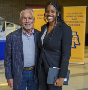 Two people smile and stand together indoors. The man on the left wears a plaid jacket and pink shirt, and the woman on the right wears a black suit and holds a tablet. Yellow college banners are visible in the background.