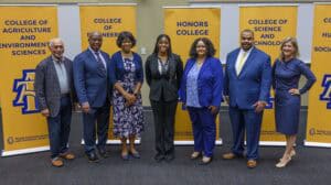 Seven professionally dressed people stand smiling in front of banners for different colleges at North Carolina A&T State University, including Agriculture and Environmental Sciences, Engineering, Honors College, and Science and Technology.
