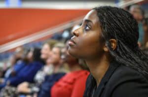 A woman with braided hair and a black blazer sits in an audience, looking attentively forward. Other people are seated beside her, slightly out of focus, in a brightly lit indoor setting.