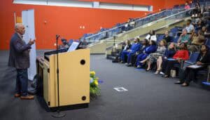 A speaker stands at a podium addressing an audience seated in a large lecture hall. The audience listens attentively, and the room features red walls and tiered seating. Flowers are arranged beside the podium.
