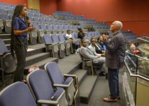A woman stands and speaks into a microphone in a mostly empty lecture hall, addressing an older man standing near the front. Several people are seated, listening attentively.