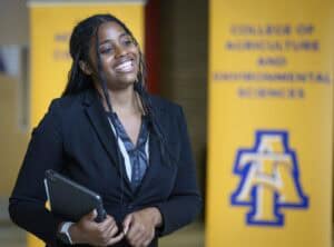 A smiling woman in a black blazer holds a tablet. She stands indoors in front of yellow banners that display text and a blue and gold logo.