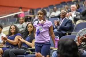 A young girl in a purple shirt stands in an auditorium while adults around her, seated in rows, applaud. She has braided hair and a plush toy around her neck. The atmosphere appears positive and supportive.
