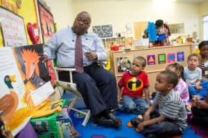A man in glasses reads a picture book to a group of young children sitting on a colorful classroom rug, with bright decorations and educational materials in the background.