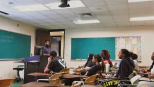 A teacher stands at a podium in front of a classroom, addressing students seated at desks. Some students have notebooks and drinks on their desks. The room has fluorescent lighting, green chalkboards, and a bulletin board.