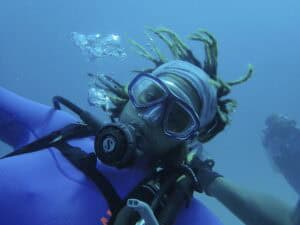 A scuba diver in a purple wetsuit and goggles looks at the camera underwater, with bubbles rising nearby and another diver visible in the background.